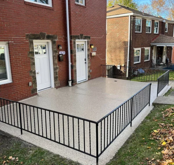 A large concrete patio with a black metal railing sits in front of a red brick duplex with identical white doors, under a clear sky.