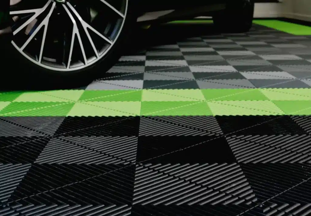 A car's wheel on a checker-patterned garage floor with black, grey, and green tiles. The tire is close-up, implying a modern, sleek vehicle.