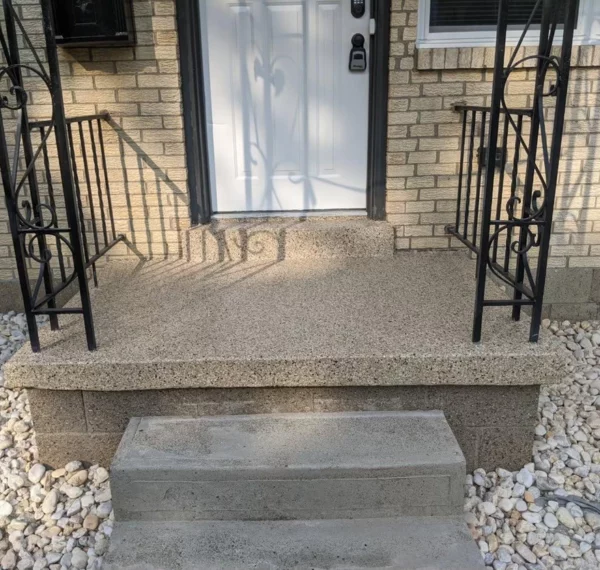The image shows a house entrance with a black door, textured steps, wrought-iron railings, and a pebbled area on either side.