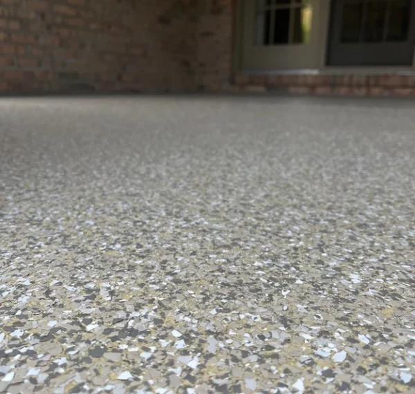 This is a close-up view of a speckled concrete surface leading to a building with visible brickwork and a glass door. The focus is on the texture.