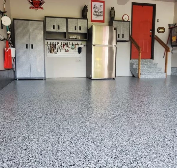 This image showcases a tidy garage interior with a speckled epoxy floor, grey cabinetry, a refrigerator, wall-mounted tool storage, and a bright red door.
