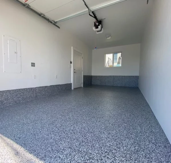 An empty garage interior with a speckled epoxy-coated floor, white walls, a window, door, ceiling-mounted garage door opener, and partial daylight.
