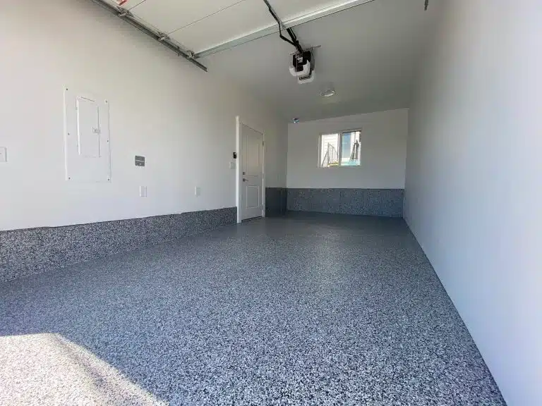 An empty garage interior with a speckled epoxy-coated floor, white walls, a window, door, ceiling-mounted garage door opener, and partial daylight.