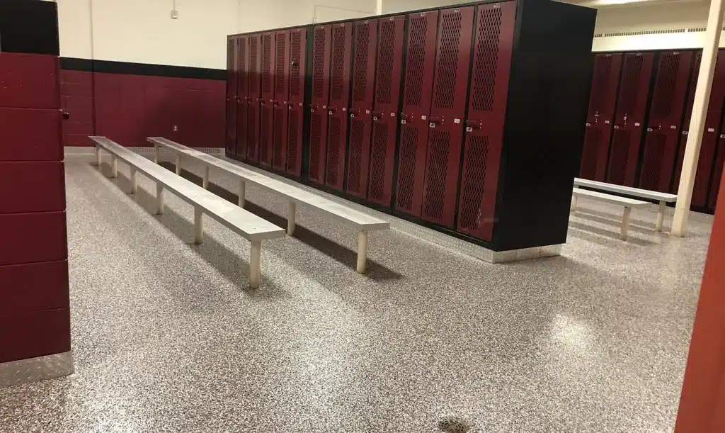 An empty locker room with red lockers lining the walls, a terrazzo floor, long benches, and a black privacy wall sectioning off an area.