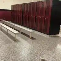 An empty locker room with red lockers lining the walls, a terrazzo floor, long benches, and a black privacy wall sectioning off an area.