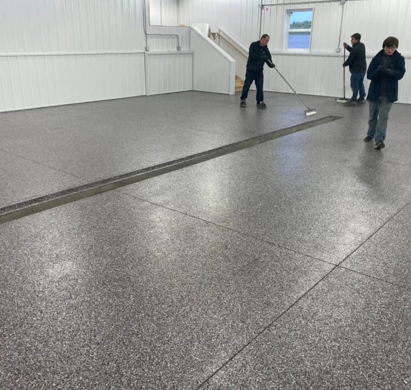 Three people in an indoor space with white walls, cleaning or working on a speckled gray floor with a squeegee and a broom.