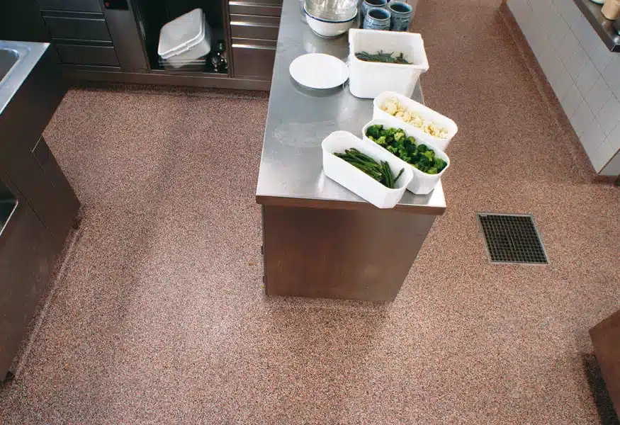 A commercial kitchen with stainless steel countertops, assorted vegetables in containers, a floor drain, and no people present. Clean and organized food prep area.