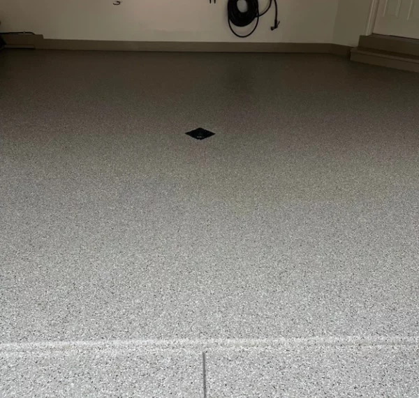 A clean, organized garage with gray speckled epoxy flooring, central floor drain, wall-mounted hose reel, and various tools neatly hung along the wall.