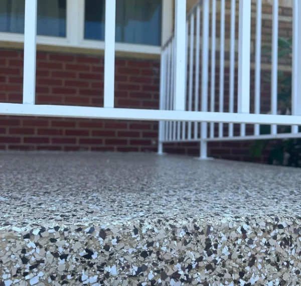 The image shows a close-up view of a textured outdoor surface leading to white metal railings, a brick wall, and windows in a residential area.
