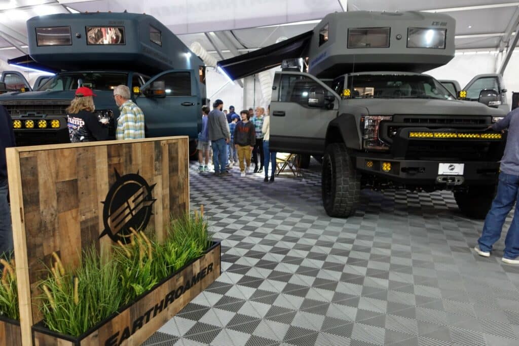 Two rugged trucks with rooftop tents are displayed at an indoor event with a checkered floor. People are examining the vehicles near a branded wooden crate.