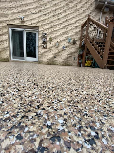 Best Patio Coating Colors An outdoor area with a textured ground leading to a brick house with a sliding door, decorative wall hangings, and wooden stairs on the side.