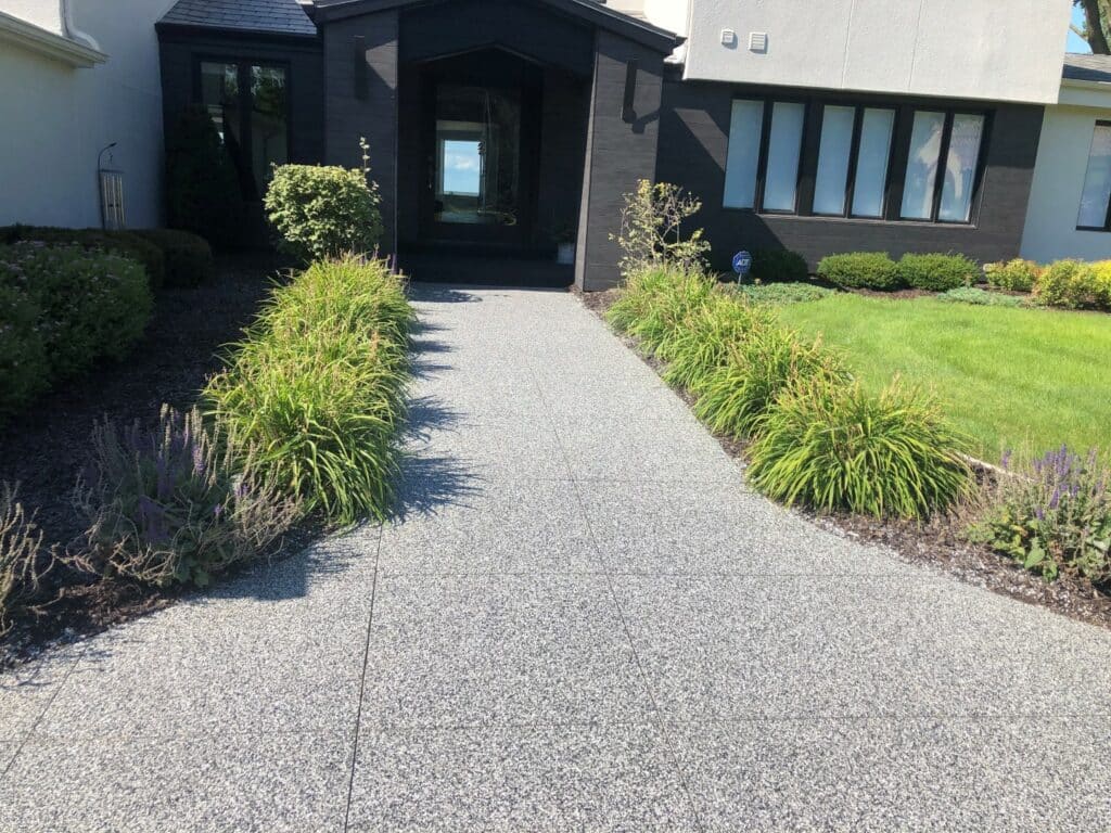 A concrete pathway leads to a black front door of a modern house with neatly landscaped grass, plants, and shrubs on a sunny day.