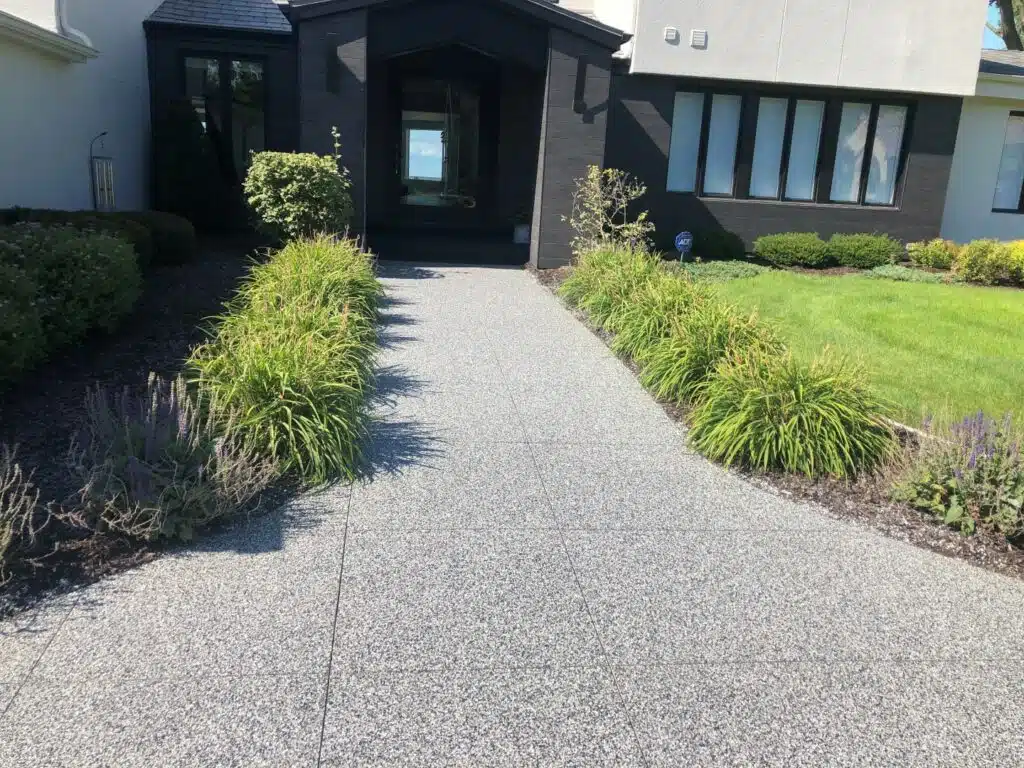 A concrete pathway leads to a black front door of a modern house with neatly landscaped grass, plants, and shrubs on a sunny day. residential concrete coatings