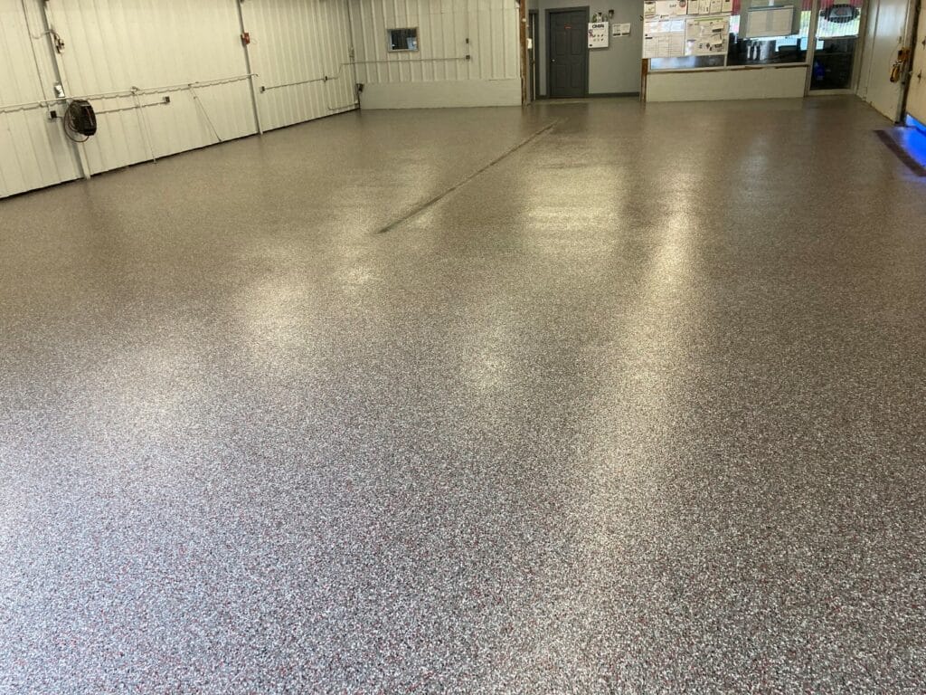 This is an image of a spacious indoor area with a shiny, speckled epoxy floor. The room contains garage doors and notice boards on distant walls.