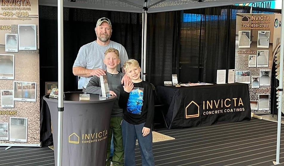 Three people stand in an exhibition booth for INVICTA Concrete Coatings, featuring various concrete samples. Two children are alongside an adult.