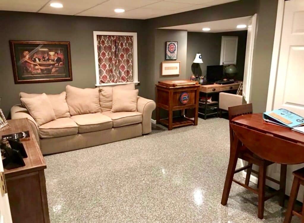 A cozy basement room with a beige couch, an office area, artwork, sports memorabilia, a table with books, and a speckled gray floor.