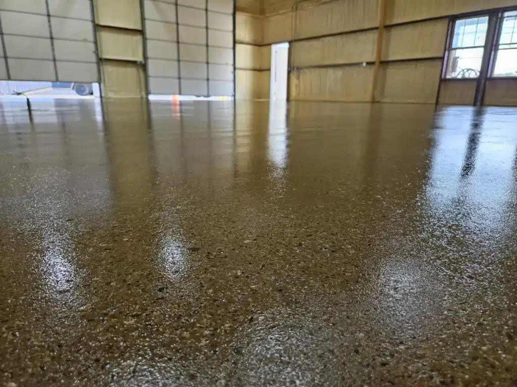 The image shows a close-up view of a shiny, polished concrete floor inside a large building with metal walls, reflecting subtle light, and a window visible.