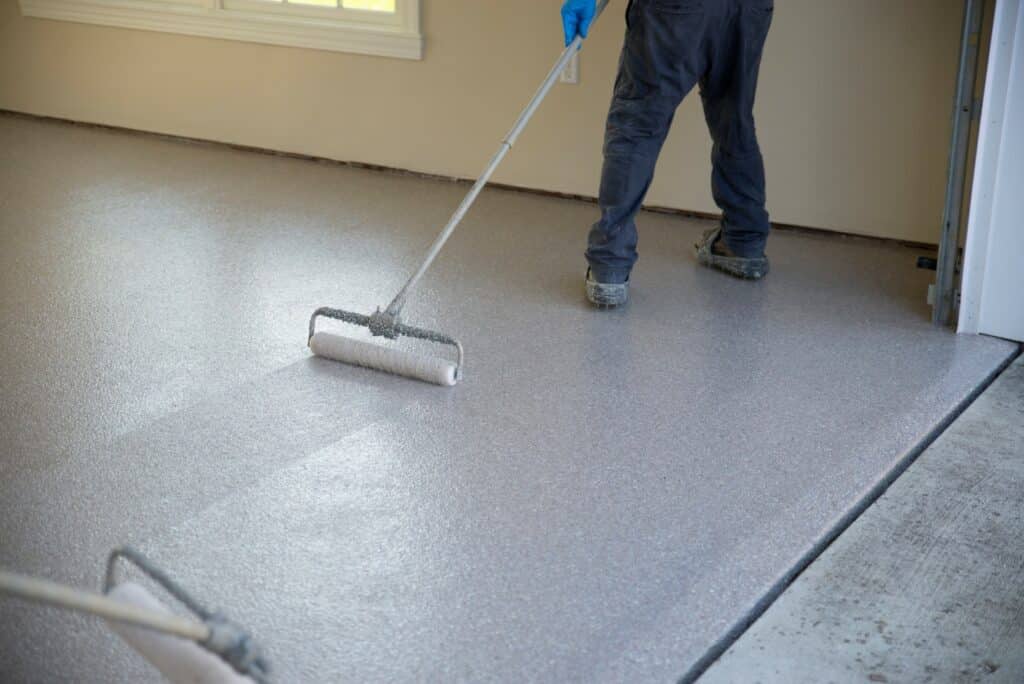 A person is applying a grey coating to a concrete floor with a roller, likely a sealant or paint, in a room with natural light coming from the left.