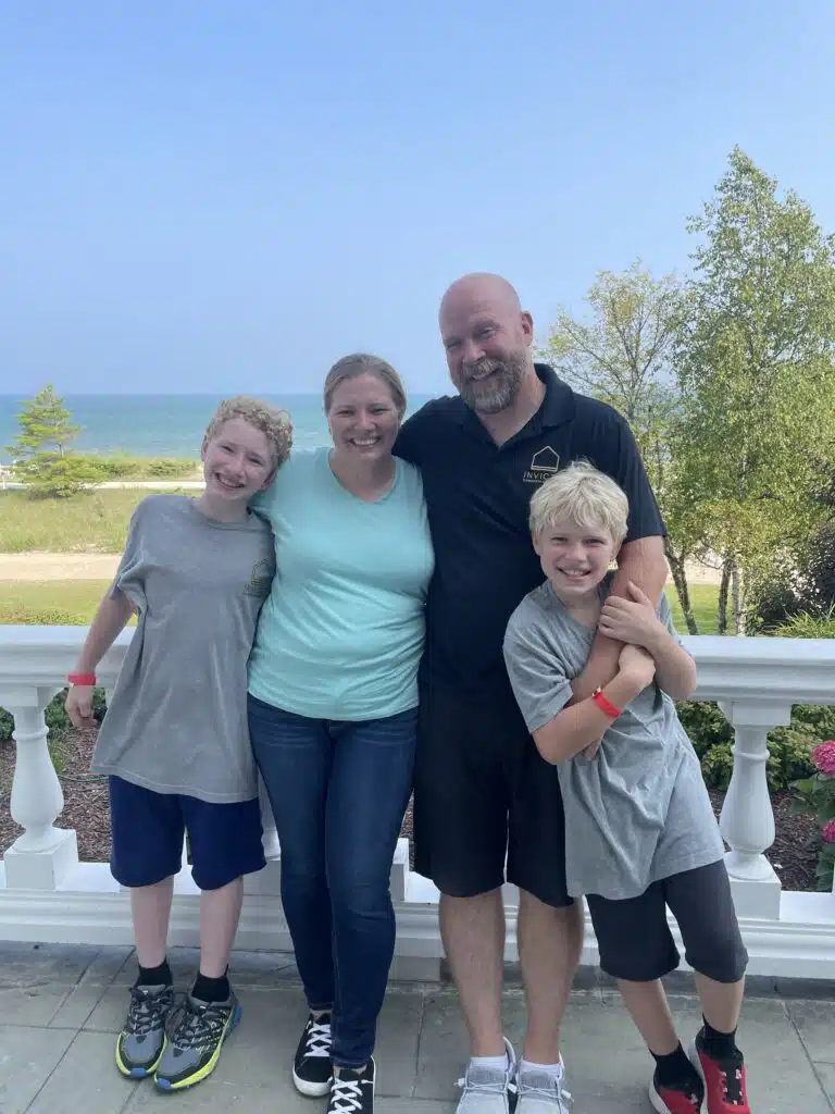 A family of four is smiling on a balcony with a beach and sea in the background, under a clear sky. Two adults and two children appear happy.