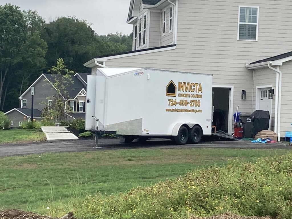 The image shows a white enclosed trailer with logos and contact information parked next to a driveway of a two-story house with green grass surroundings.