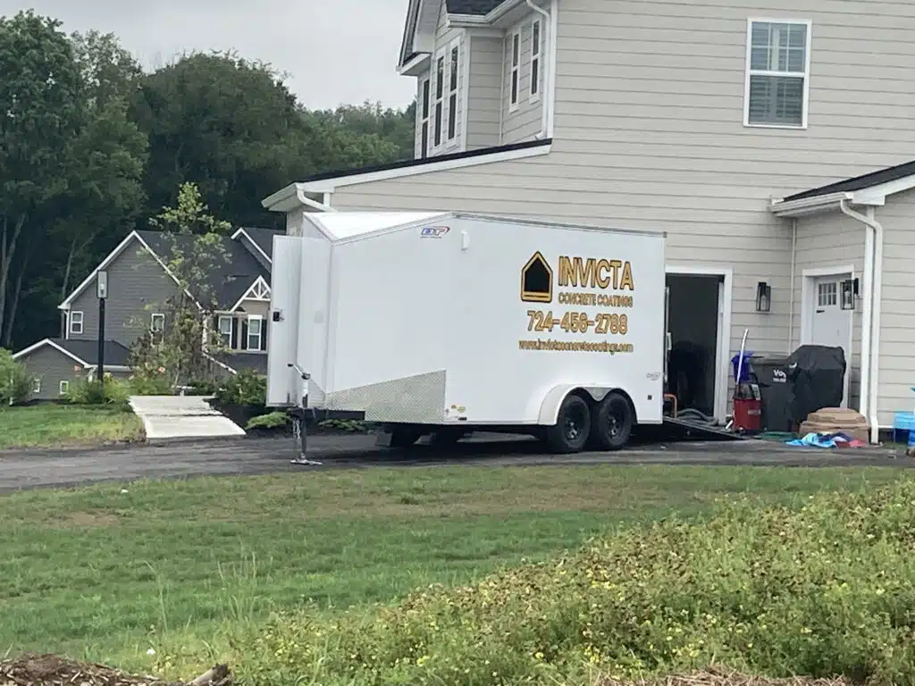 The image shows a white enclosed trailer with logos and contact information parked next to a driveway of a two-story house with green grass surroundings. about