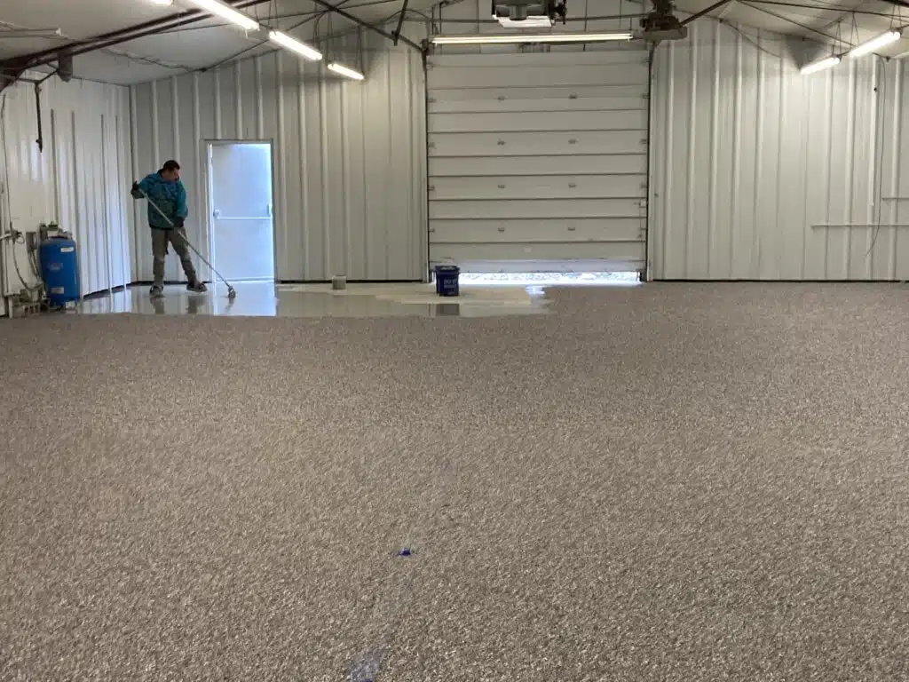 A person is standing inside a spacious, brightly-lit garage with a speckled floor, leaning on a broom near the open entrance, with cleaning equipment nearby.