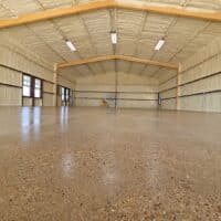This image shows a spacious, empty indoor hall with a polished concrete floor, timber framing, beige walls, large windows, and a yellow ladder in the distance.