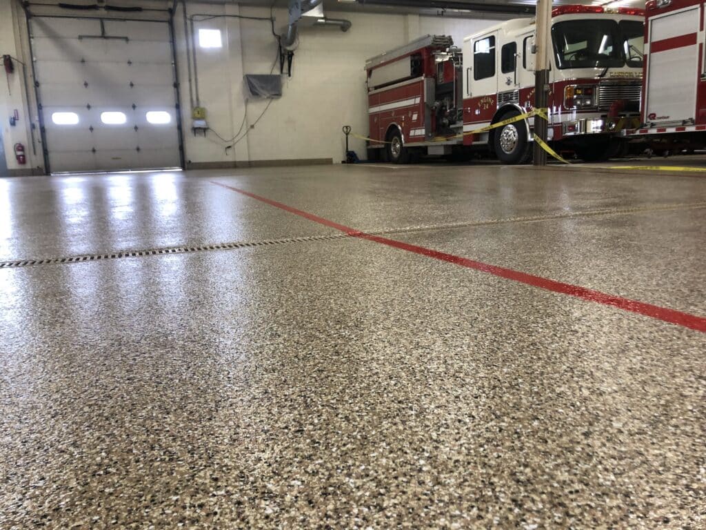 This image shows a close-up of a textured floor inside a fire station, with a firetruck parked in the background, next to an open garage door.