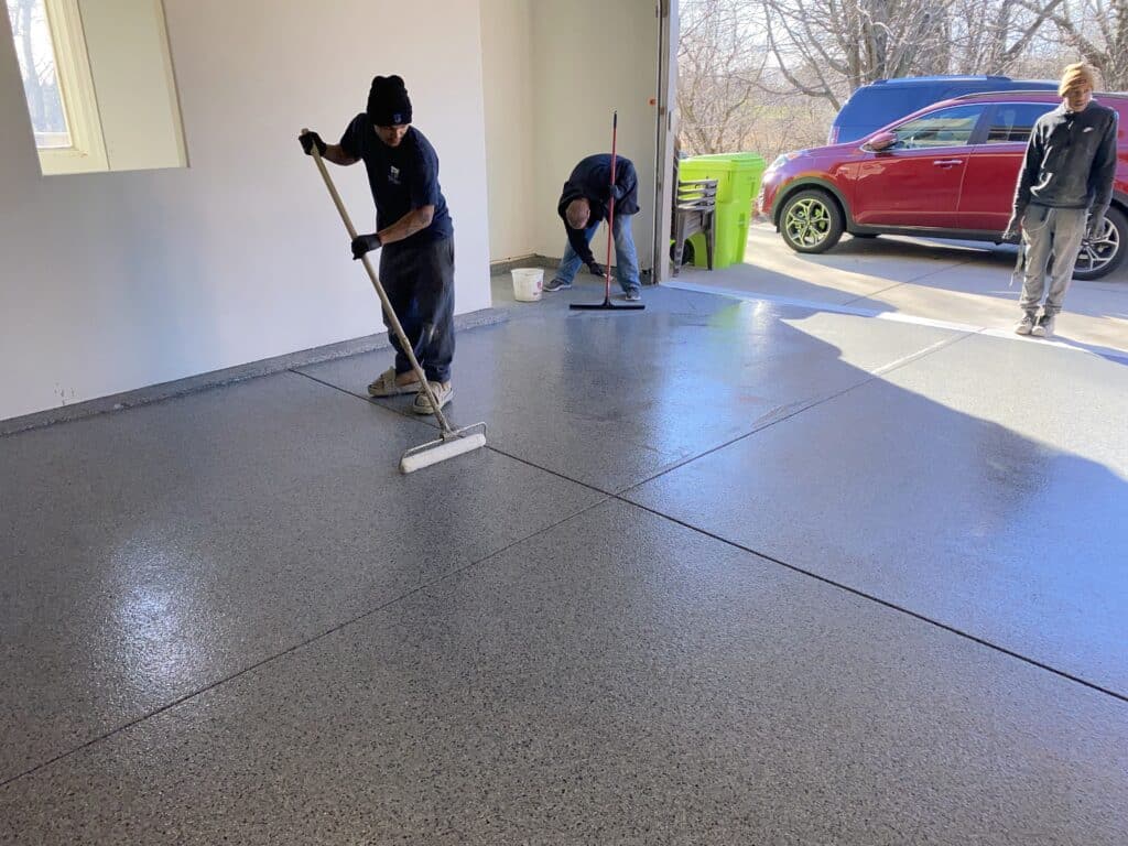 Two people are applying a coating to a garage floor. Another person observes. A red vehicle is visible outside. The environment appears clean and bright.