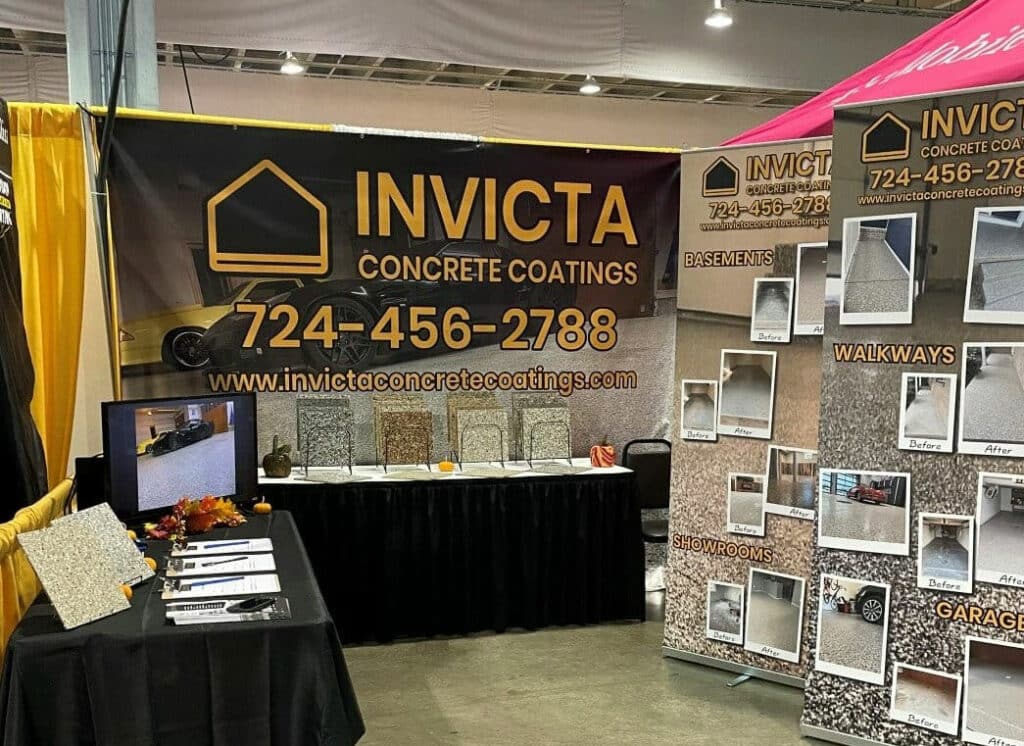 The image shows an exhibition booth for INVICTA Concrete Coatings with sample displays, a flat-screen TV, informational posters, and a black draped table.