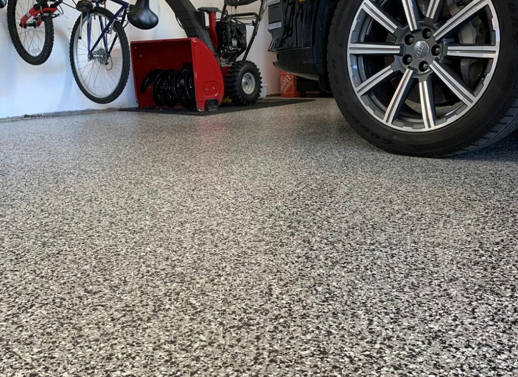 The image shows a close-up view of a speckled epoxy garage floor. Part of a car's tire, a bicycle, and red machinery are visible on the sides.