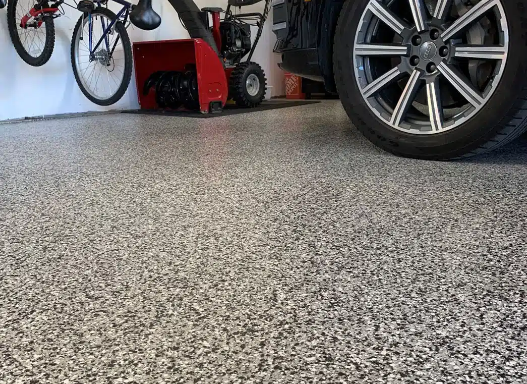 The image shows a close-up view of a speckled epoxy garage floor. Part of a car's tire, a bicycle, and red machinery are visible on the sides. concrete coatings near me polyurea garage floor coating cost