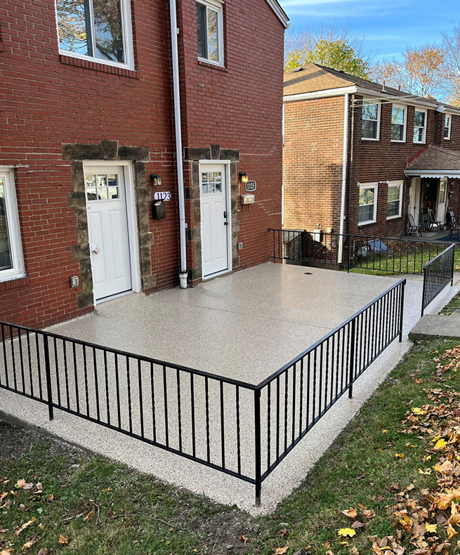 The image shows a red brick duplex with two white doors, each with a black number plaque. A freshly coated concrete patio with a black railing is in front.