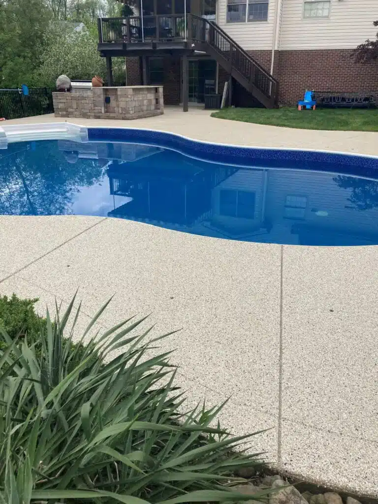 This image depicts a serene backyard with an inground pool, surrounding concrete deck, green foliage, and a house with a raised deck where a person is sitting.