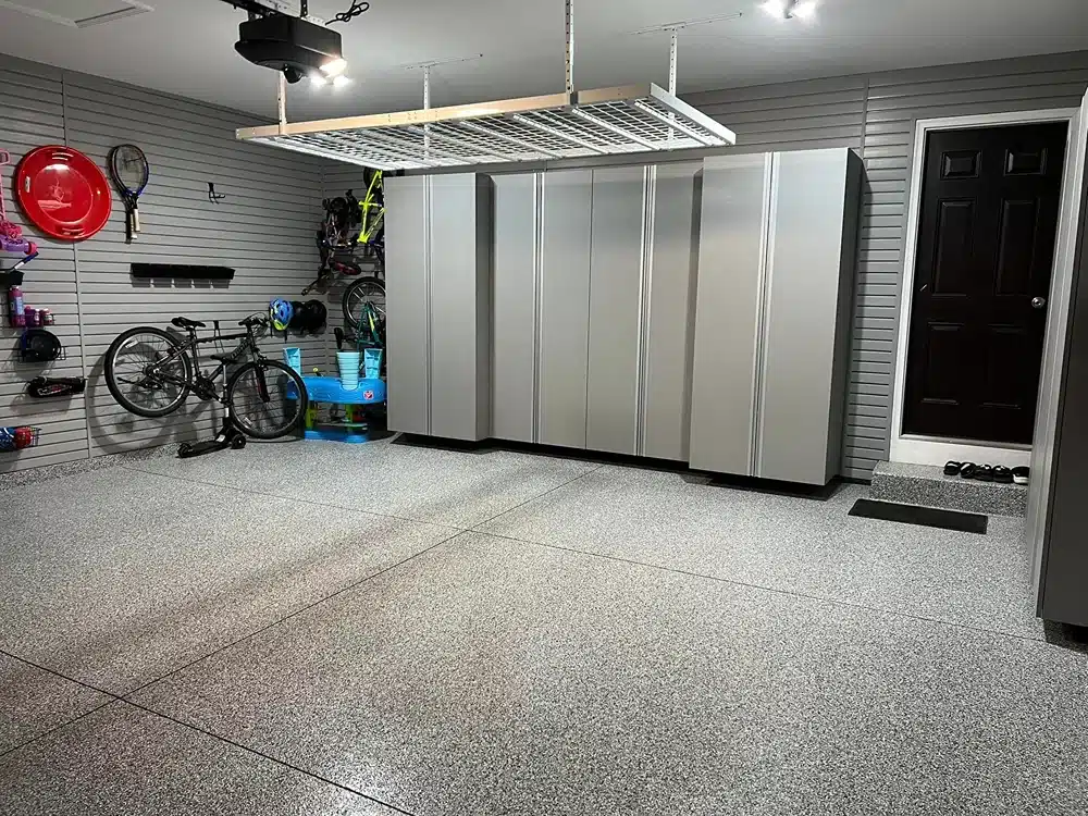 Organized garage interior with epoxy flooring, gray storage cabinets, bicycles, sports equipment, a dark entry door, and a ceiling storage rack.