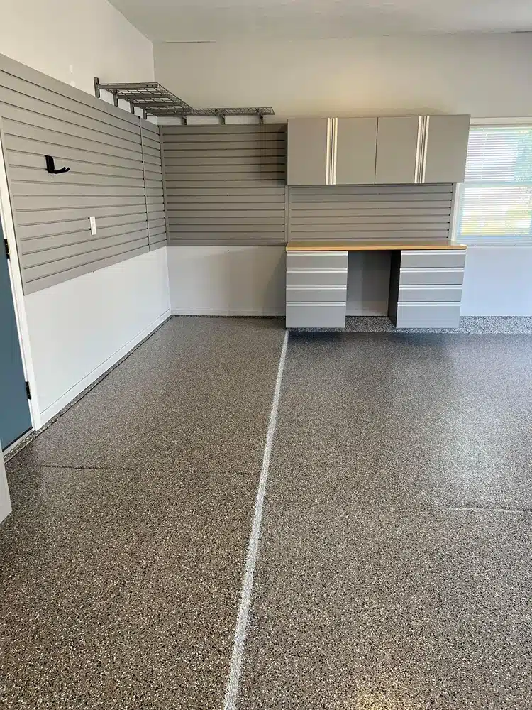 The image shows a tidy garage interior with a speckled floor, gray storage cabinets, slatwall panels for tools, and a workbench under a window.