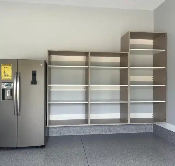 A stainless steel refrigerator stands next to empty wooden bookshelves in a room with a grey floor and a two-tone painted wall.