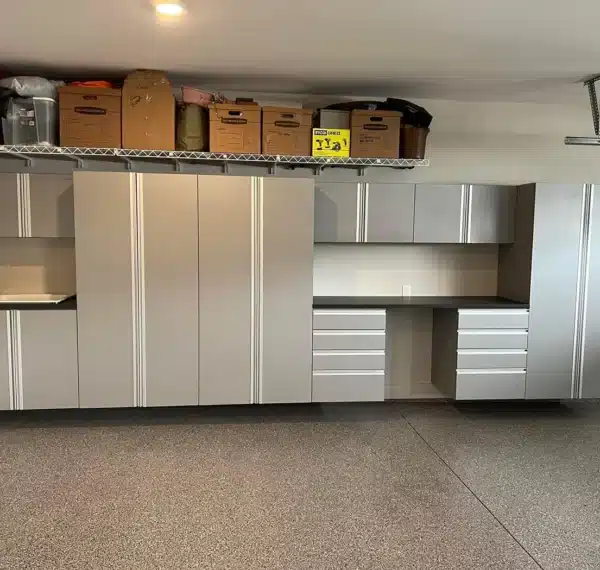 The image shows a neat garage interior with gray storage cabinets, a workbench, a speckled floor, and various boxes stored on overhead shelving.