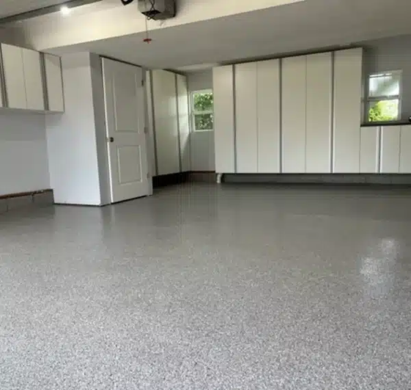 The image shows an empty garage interior with white cabinets, a sealed gray floor, a closed door, and natural light coming from a window.
